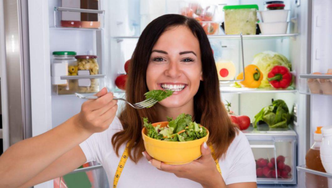 femeie care sta cu frigiderul deschis si mananca dintr-un bol salata zambind