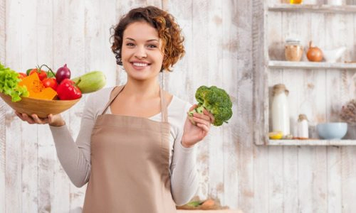 femeie ce zambete intr-o bucatarie care tine in mana stanga brocoli iar in dreapta un bol cu legume