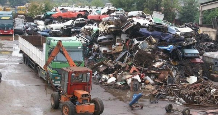 Un tractor portocaliu si un TIR verde, langa un morman de masini in proces de reciclare