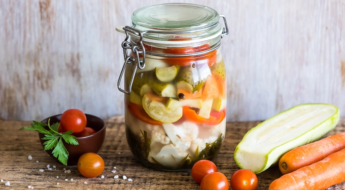 un borcan de muraturi asortate langa mai multe legume proaspete pe o masa