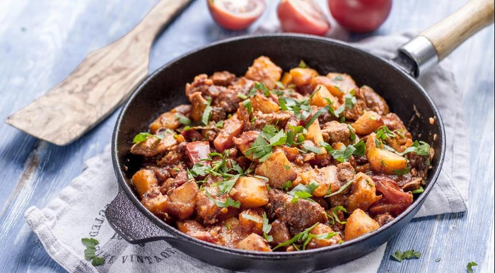 friptura de porc cu cartofi intr-un bol langa o lingura din lemn si rosii 