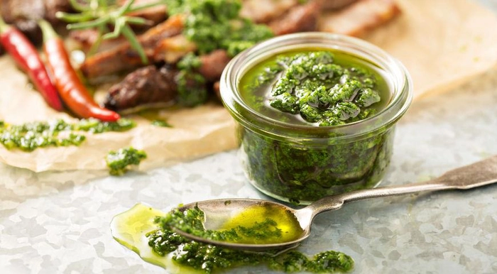 o lingura cu sos chimichurri langa un bol cu sos si carne pe o masa