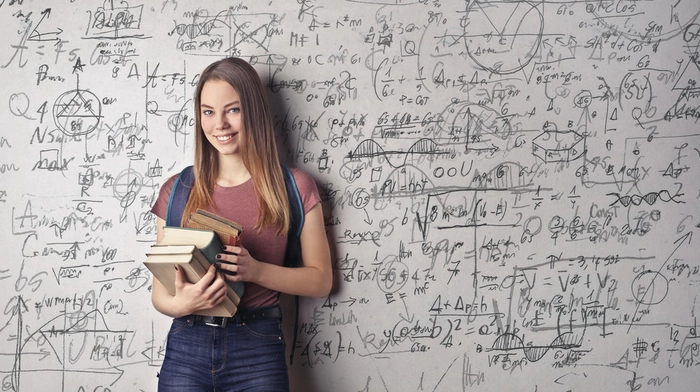 femeie care sta cu mai multe carti in mana langa o tabla plina de formule matematice si zambeste
