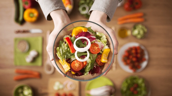 persoana care tine in mana un bol cu salata deasupra unei mese pline cu alimente sanatoase