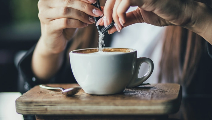 maini ce adauga zahar intr-o ceasca de cafea, lingurita