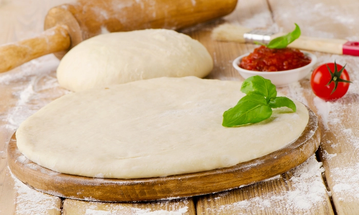 un blat pentru pizza pe un platou din lemn langa aluat si alte ingrediente