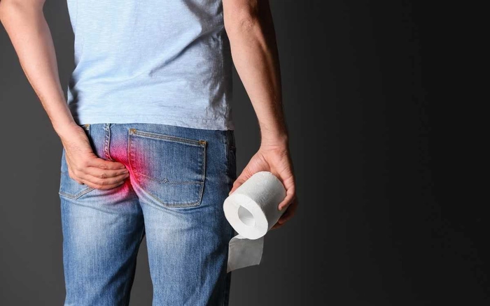 grafica barbat care se tine de fund din cauza disconfortului provocat de hemoroizi si tine o hartie igienica in mana