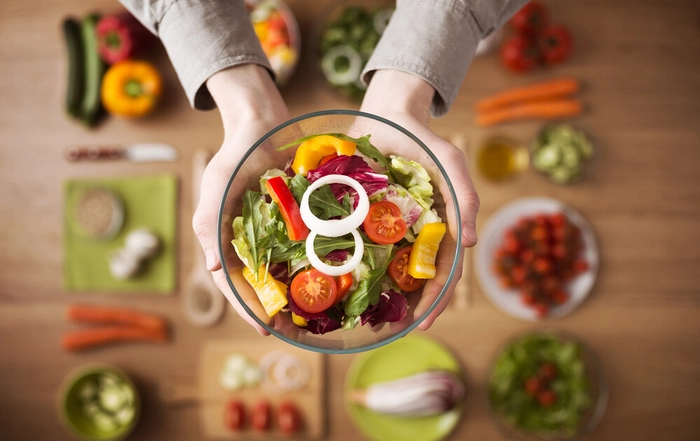 persoana care tine un bol cu salata in mana deasupra unei mese plina cu diverse ingrediente de post