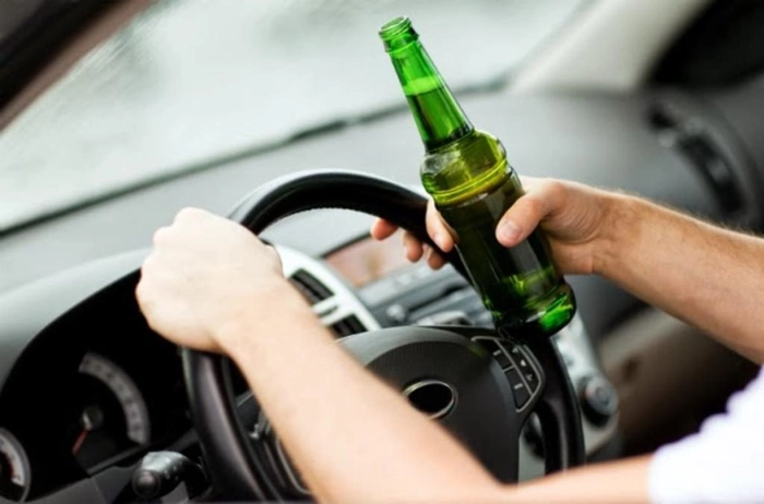 Mainile unui barbat pe volanul unei masini; o sticla ce contine alcool, in mana dreapta a soferului
