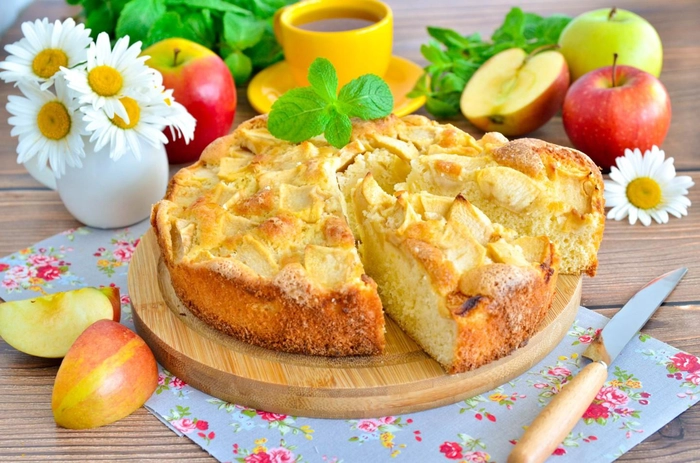 o prajitura turnata taiata cu mere pe o farfurie langa o cana cu ceai si mai multe fructe proaspete