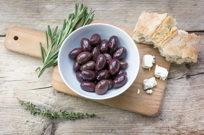 un bol cu masline kalamata pe o masa langa mai multe condimente si paine
