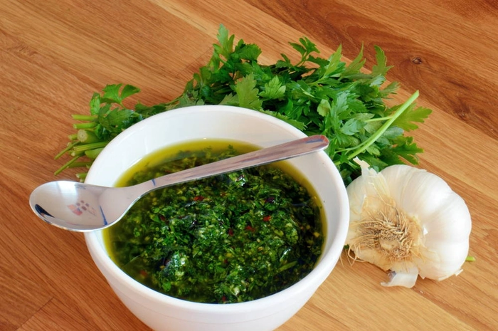 o lingura pe un bol cu sos chimichurri langa un catel de usturoi si patrunjel