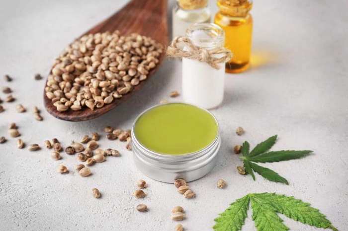 o lingura de lemn cu seminte de canepa langa un recipient cu ulei de canepa si o crema