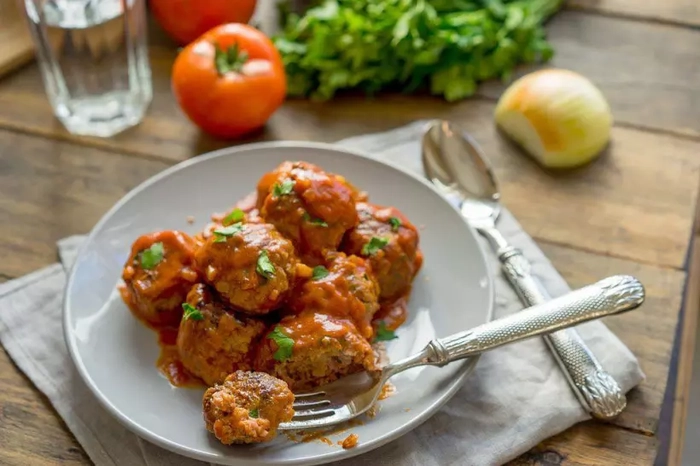 mai multe chiftelute in sos de rosii pe o farfurie langa mai multe ingrediente si tacamuri