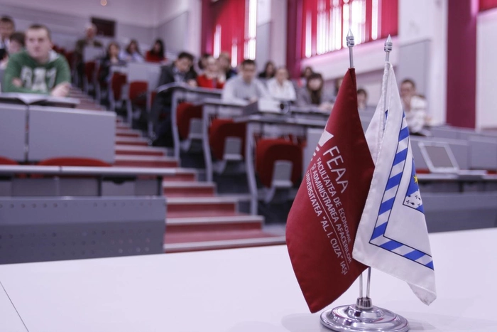 studenti intr-un amfiteatru, iar în prim plan sunt doua steaguri cu feea si cu sigla universitatii