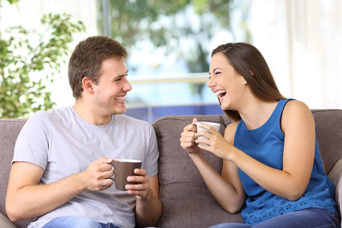 o femeie si un barbat care stau asezati pe canapea si beau cate o cafea zambind