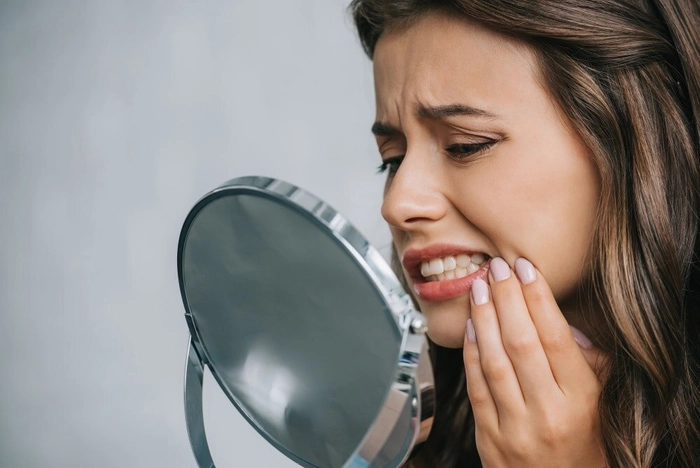 femeie care se tine de obraz din cauza durerii simtite la nivelul gingiilor si priveste in oglinda