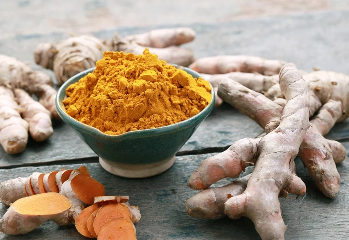 un bol cu praf de turmeric langa mai multe radacini de curcuma pe o masa