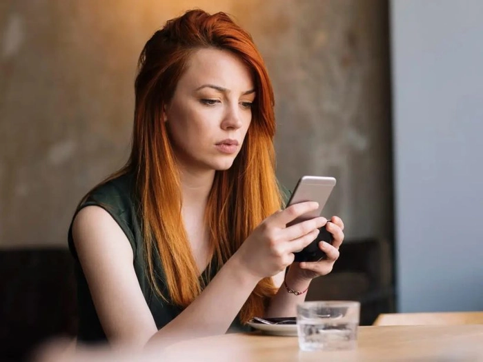  O femeie foloseste un smartphone, sta la o masa crem, pe care este un pahar de sticla