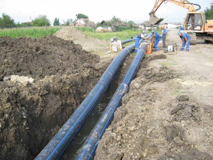 muncitori ce pun tevi mari in pamant, in fundal un excavator