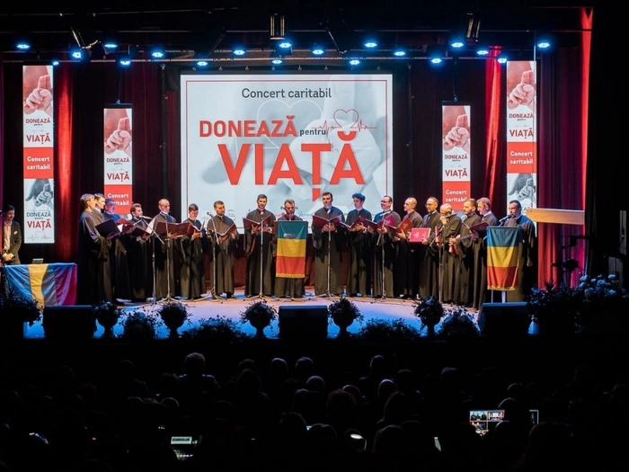 Un cor de preoti cantand pe o scena cu steaguri in mana,iar pe ecran fiind afisata o imagine pentru o campanie de caritate