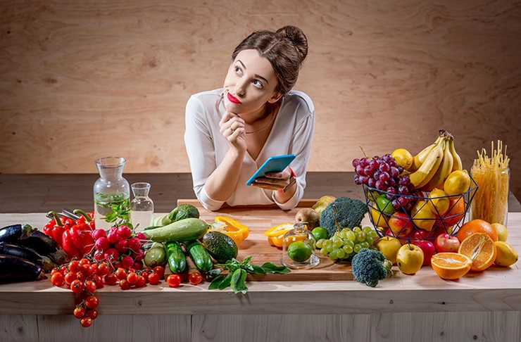 femeie care sta cu un telefon in mana si se gandeste fiind inconjurata cu mai multe fructe si legume