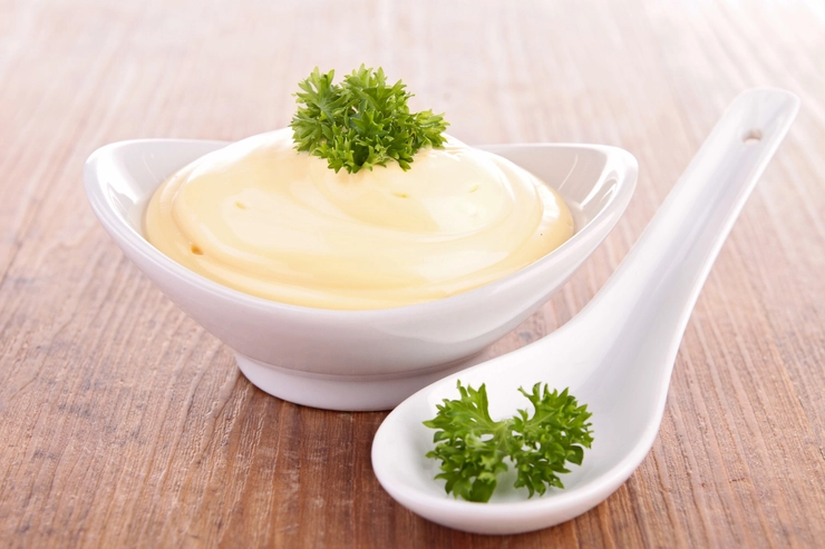 un bol cu sos aioli decorat cu verdeata pe o masa alaturi de o lingura pentru sos