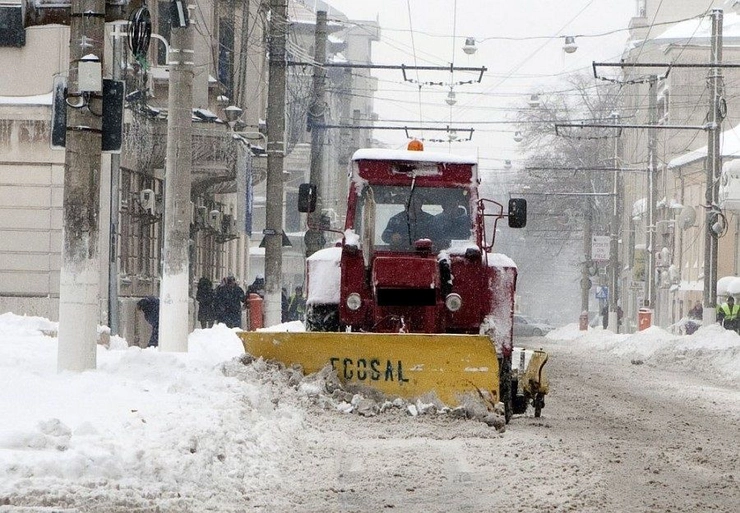 un automobil de deszapezire in timpul functionarii, pe o strada cu zapada si cu persoane pe trotuare in plan secund